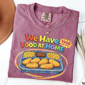 May include: Berry-colored t-shirt with the text "We Have Food At Home 1998 Edition" and a graphic of a TV dinner. A gold necklace and a brown hat are also visible.