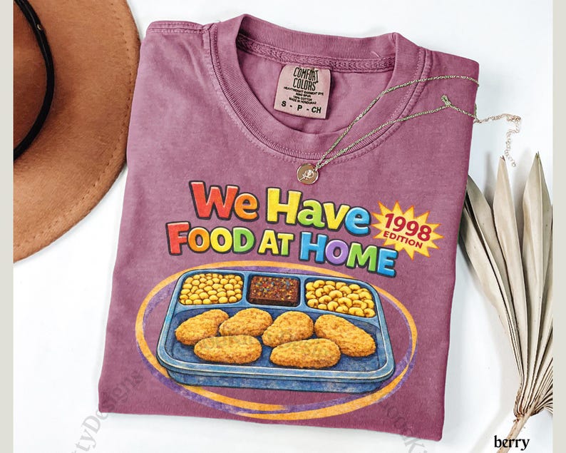 May include: Berry-colored t-shirt with the text "We Have Food At Home 1998 Edition" and a graphic of a TV dinner. A gold necklace and a brown hat are also visible.