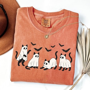 May include: A yam-colored t-shirt featuring a graphic of four cats dressed as ghosts with black bat silhouettes. The shirt has a crew neck and a small tag on the upper left. A gold necklace and a brown hat are also visible.