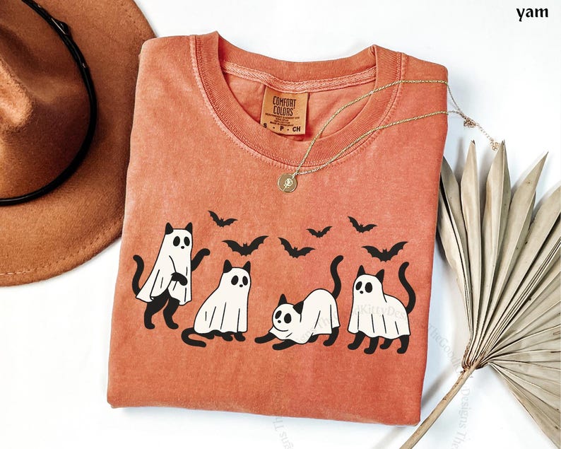 May include: A yam-colored t-shirt featuring a graphic of four cats dressed as ghosts with black bat silhouettes. The shirt has a crew neck and a small tag on the upper left. A gold necklace and a brown hat are also visible.