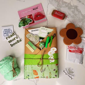 May include: A brown paper gift bag with green and white floral wrapping paper. The bag is tied with twine and decorated with stickers that say "Reading List" and "Books of". The bag is surrounded by other items, including a green washcloth, a tea bag, a chocolate packet, a cookie, and a lip balm.