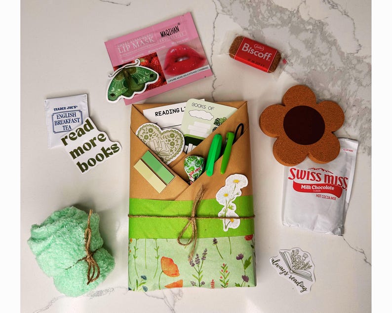 May include: A brown paper gift bag with green and white floral wrapping paper. The bag is tied with twine and decorated with stickers that say "Reading List" and "Books of". The bag is surrounded by other items, including a green washcloth, a tea bag, a chocolate packet, a cookie, and a lip balm.