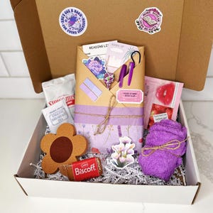 May include: A white box filled with a bookish themed gift set. The box contains a purple towel, a brown flower-shaped coaster, a book-shaped sticker, a purple and white patterned journal, a package of tea, a pink lip balm, a package of cookies, and a purple and white patterned envelope with a sticker that says "House of Wine Book Club".