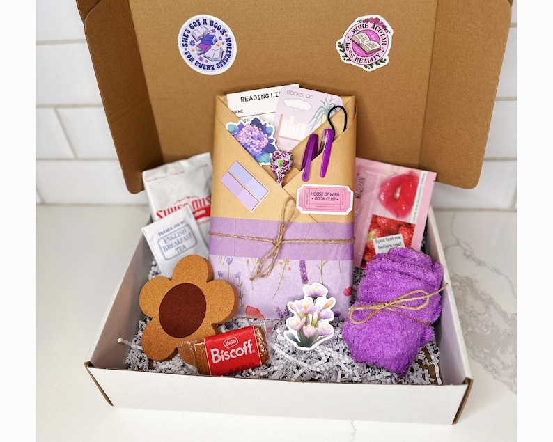 May include: A white box filled with a bookish themed gift set. The box contains a purple towel, a brown flower-shaped coaster, a book-shaped sticker, a purple and white patterned journal, a package of tea, a pink lip balm, a package of cookies, and a purple and white patterned envelope with a sticker that says "House of Wine Book Club".