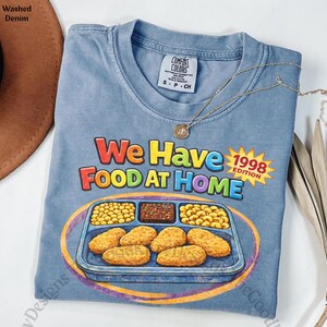 May include: A light blue t-shirt with the text "We Have Food At Home" and a graphic of a school lunch tray. The shirt also has the text "1998 Edition" in a yellow starburst. A gold necklace is draped across the shirt.