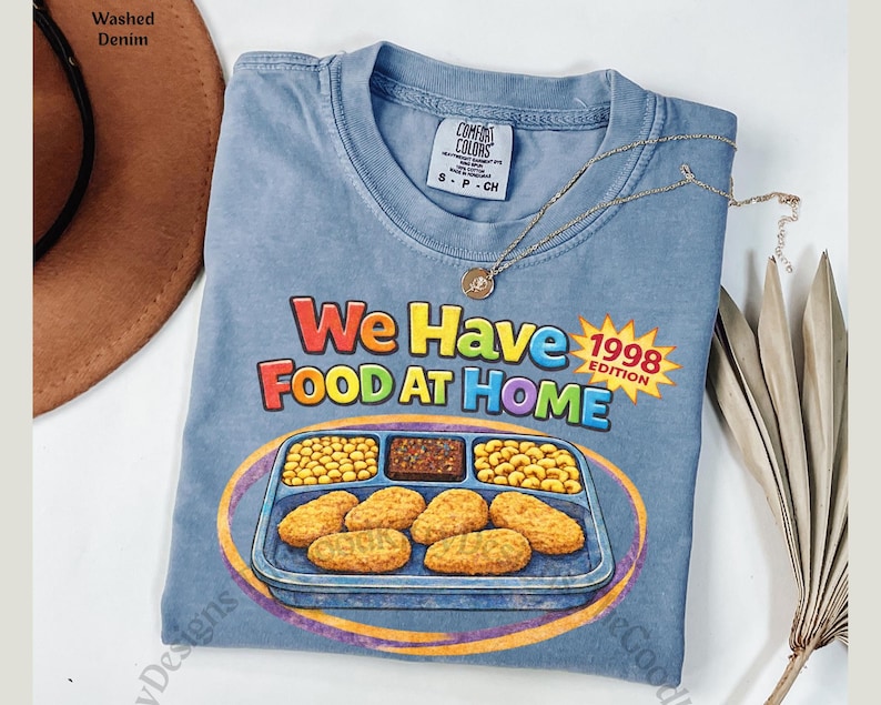 May include: A light blue t-shirt with the text "We Have Food At Home" and a graphic of a school lunch tray. The shirt also has the text "1998 Edition" in a yellow starburst. A gold necklace is draped across the shirt.