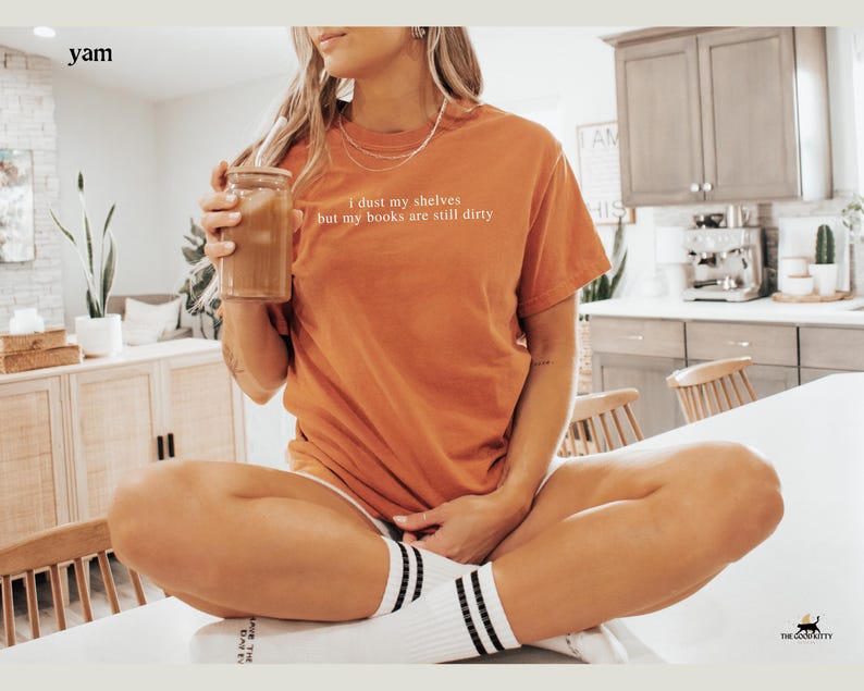 May include: A woman wearing a burnt orange t-shirt with the text "I dust my shelves but my books are still dirty" sitting on a white surface holding a glass of iced coffee.