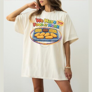 May include: Cream-colored oversized t-shirt with a colorful graphic. The graphic features a cartoon-style tray of food with the text "We Have Food At Home" and "1998 Edition". The shirt has short sleeves and a relaxed fit.