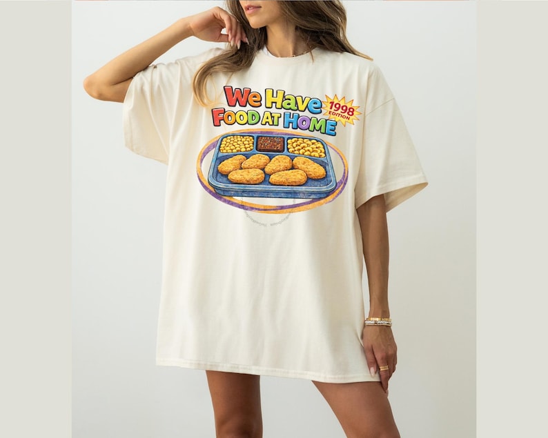 May include: Cream-colored oversized t-shirt with a colorful graphic. The graphic features a cartoon-style tray of food with the text "We Have Food At Home" and "1998 Edition". The shirt has short sleeves and a relaxed fit.