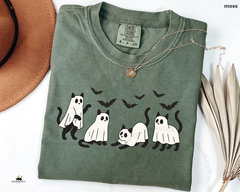 May include: Moss green t-shirt featuring a graphic of four cats dressed as ghosts with black bat silhouettes above them. The shirt has a crew neck and short sleeves. The brand label is visible on the upper left. A gold necklace is draped across the shirt.
