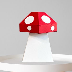 Puede incluir: Una seta de papel hecha a mano, con un sombrero rojo y lunares blancos. La seta está sobre un tallo blanco, colocada sobre una mesa auxiliar redonda blanca con patas de madera. El fondo es gris claro liso.