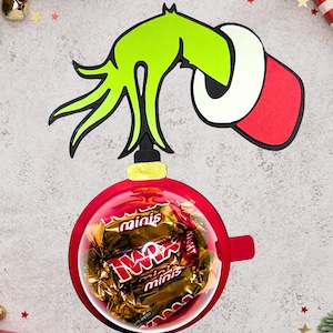 Puede incluir: Una mano verde del Grinch sosteniendo un adorno navideño rojo lleno de mini barritas de chocolate. El adorno está rodeado de estrellas doradas y cinta roja y dorada.