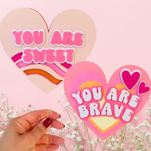 Puede incluir: Dos corazones de papel rosa con diseños de papel en capas. El corazón superior tiene el texto "You are sweet" en letras rosas. El corazón inferior tiene el texto "You are brave" en letras blancas.