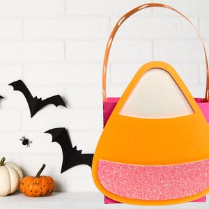 Puede incluir: Una bolsa de golosinas con forma de caramelo de Halloween con un acento de purpurina rosa. La bolsa tiene un cuerpo naranja, un interior blanco y una base rosa. Murciélagos de papel negro y calabazas decoran el fondo.