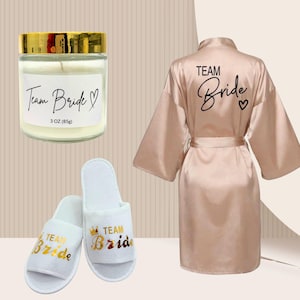 Puede incluir: Una vela de 85g en un frasco de vidrio con tapa dorada, un par de pantuflas blancas abiertas y una bata de satén color champán. La etiqueta de la vela, las pantuflas y la parte posterior de la bata muestran el texto Team Bride con un corazón.