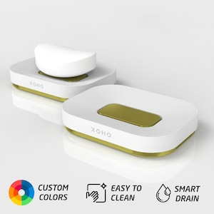 May include: White soap dish with a gold trim and the word "XOHO" printed on the front. The dish has a built-in drainage system. The soap dish is shown with a white bar of soap on top.