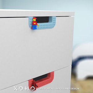 May include: White dresser with two drawers. The top drawer has a blue Lego-style handle with a red Lego figure. The bottom drawer has a red Lego-style handle with a red Lego figure. The text "XOXO" is visible at the bottom of the image.