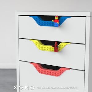 May include: A white dresser with three drawers. Each drawer has a different colored Lego handle: blue, yellow, and red. The Lego handles are shaped like a curved line.