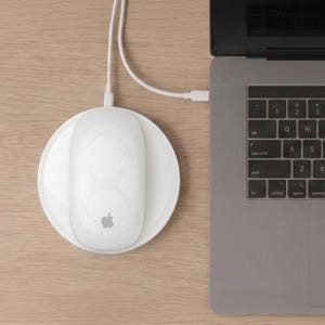 May include: A white wireless computer mouse with a charging base, connected to a laptop via a white cable. The mouse has a smooth surface with a subtle design and the Apple logo. The laptop is silver and has a keyboard.