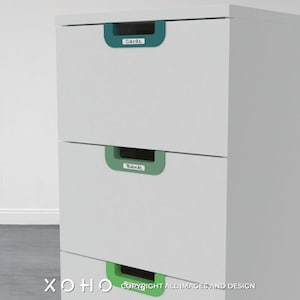 Könnte beinhalten: Weiße Kommode mit drei Schubladen. Jede Schublade hat einen grünen oder türkisfarbenen Griff mit einem handgeschriebenen Etikett. Die oberste Schublade ist mit "Cards" beschriftet, die mittlere Schublade mit "Bank" und die unterste Schublade ist unbeschriftet.