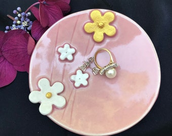 May include: A pink ceramic trinket dish with white and gold flower designs. The dish is decorated with three white flowers with gold centers and one larger gold flower. A ring and earrings are displayed on the dish.