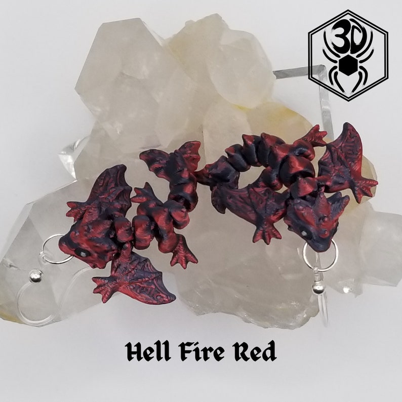 K&ouml;nnte beinhalten: Ein Paar rote und schwarze Drachenohrringe mit silbernen Haken. Die Drachen sind zusammengerollt mit ausgebreiteten Fl&uuml;geln. Die Ohrringe sind mit "Hell Fire Red" beschriftet.