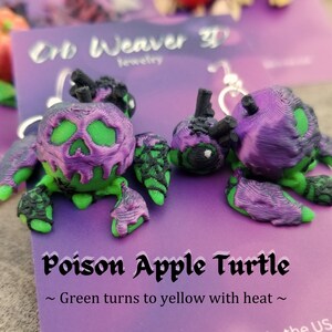 K&ouml;nnte beinhalten: Ein Paar gr&uuml;ne und violette Ohrringe in Form einer vergifteten Apfel-Schildkr&ouml;te. Die Ohrringe sind aus 3D-gedrucktem Kunststoff gefertigt und haben ein Totenkopfdesign auf dem Apfel. Der Text auf der Karte lautet "Poison Apple Turtle ~ Green turns to yellow with heat ~".