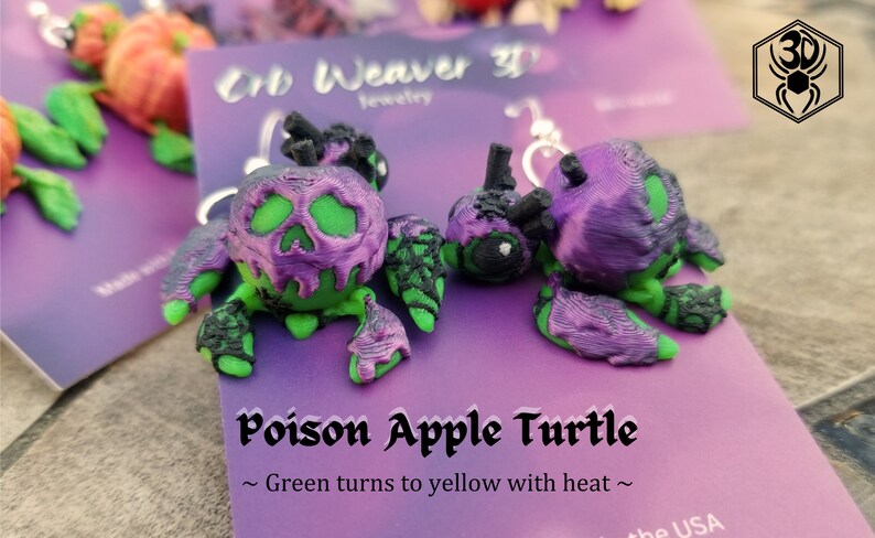 K&ouml;nnte beinhalten: Ein Paar gr&uuml;ne und violette Ohrringe in Form einer vergifteten Apfel-Schildkr&ouml;te. Die Ohrringe sind aus 3D-gedrucktem Kunststoff gefertigt und haben ein Totenkopfdesign auf dem Apfel. Der Text auf der Karte lautet "Poison Apple Turtle ~ Green turns to yellow with heat ~".