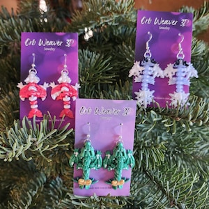 May include: Three pairs of handmade earrings on purple cards, displayed on a bed of evergreen branches. The earrings feature unique designs: red and white figures, white and black figures, and green dragon-like creatures. Each card reads "Crb Weaver 3D Jewelry."
