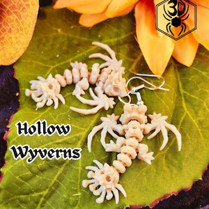 May include: A pair of white skeletal wyvern earrings on a green leaf background. The earrings are made of polymer clay and have a hollow, detailed design. The text "Hollow Wyverns" is visible in the image.