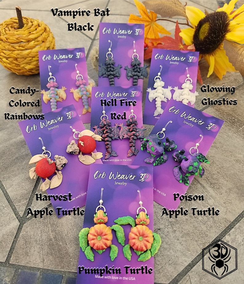 K&ouml;nnte beinhalten: Eine Auswahl handgefertigter Ohrringe aus Polymer Clay mit verschiedenen Halloween- und Herbstmotiven. Die Ohrringe werden auf violetten Karten mit dem Text "Orb Weaver 3D Jewelry" und den Namen der jeweiligen Designs pr&auml;sentiert: "Vampire Bat Black", "Candy-Colored Rainbows", "Hell Fire Red", "Glowing Ghosties", "Harvest Apple Turtle", "Poison Apple Turtle" und "Pumpkin Turtle".