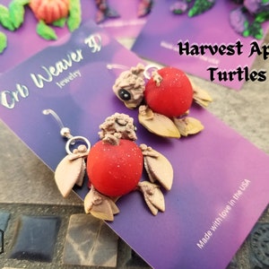 K&ouml;nnte beinhalten: Ein Paar handgefertigte Ohrringe aus Polymer Clay, die eine Schildkr&ouml;te mit einer roten Apfelschale zeigen. Die Ohrringe sind mit "Harvest Apple Turtles" und "Orb Weaver 3D Jewelry" beschriftet.