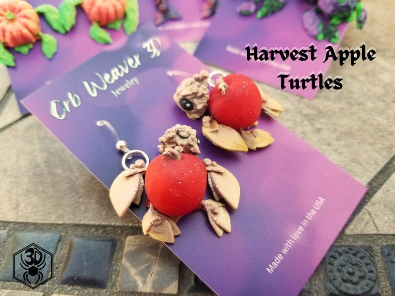 K&ouml;nnte beinhalten: Ein Paar handgefertigte Ohrringe aus Polymer Clay, die eine Schildkr&ouml;te mit einer roten Apfelschale zeigen. Die Ohrringe sind mit "Harvest Apple Turtles" und "Orb Weaver 3D Jewelry" beschriftet.