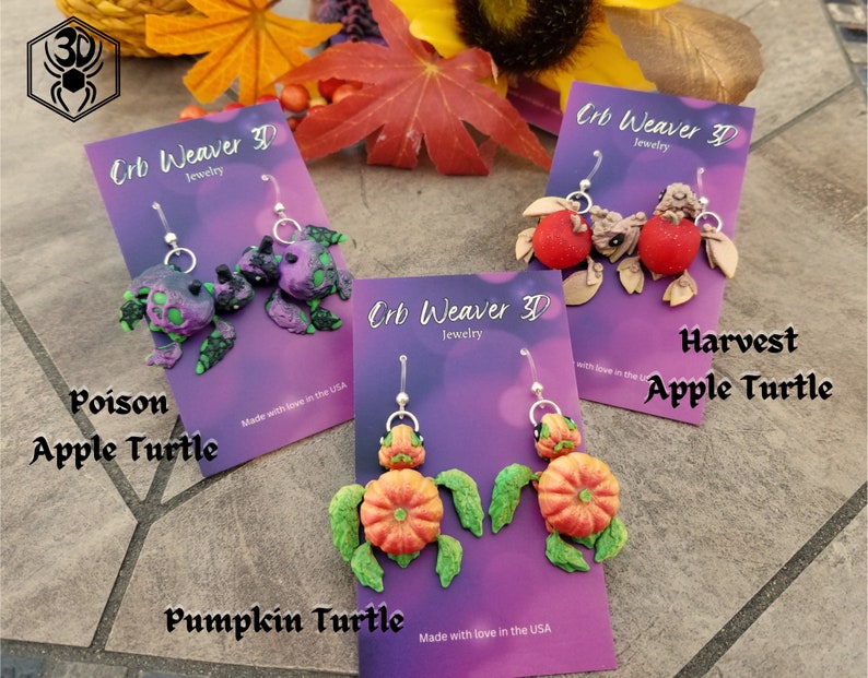 K&ouml;nnte beinhalten: Vier Paar handgefertigte Ohrringe aus Polymer Clay mit einem Schildkr&ouml;ten-Design. Die Ohrringe sind in vier verschiedenen Stilen erh&auml;ltlich: Poison Apple Turtle, Harvest Apple Turtle, Pumpkin Turtle und eine gr&uuml;ne und violette Schildkr&ouml;te. Jedes Paar Ohrringe ist mit dem Namen des Stils und dem Markennamen "Crb Weaver 3D Jewelry" gekennzeichnet.