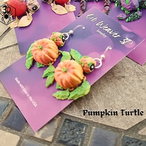 May include: A pair of pumpkin turtle earrings with green leaves. The earrings are made of polymer clay and are handmade. The earrings are on a purple card with the text "Oh Weaver D Jewelry" and "Pumpkin Turtle".