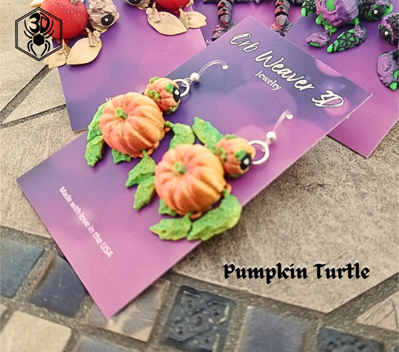 K&ouml;nnte beinhalten: Ein Paar K&uuml;rbis-Schildkr&ouml;ten-Ohrringe mit gr&uuml;nen Bl&auml;ttern. Die Ohrringe sind aus Polymer Clay gefertigt und handgefertigt. Die Ohrringe befinden sich auf einer lila Karte mit dem Text "Oh Weaver D Jewelry" und "Pumpkin Turtle".