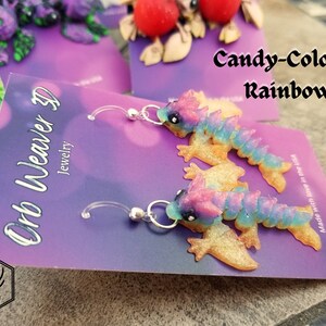 K&ouml;nnte beinhalten: Ein Paar H&auml;ngeohrringe mit einem verspielten, bonbonfarbenen Drachen-Design. Der Drache ist aus Polymer Clay gefertigt und hat eine glitzernde Oberfl&auml;che. Die Ohrringe befinden sich auf einer violetten Karte mit dem Text "Orb Weaver 3D Jewelry" und "Candy-Colored Rainbow".