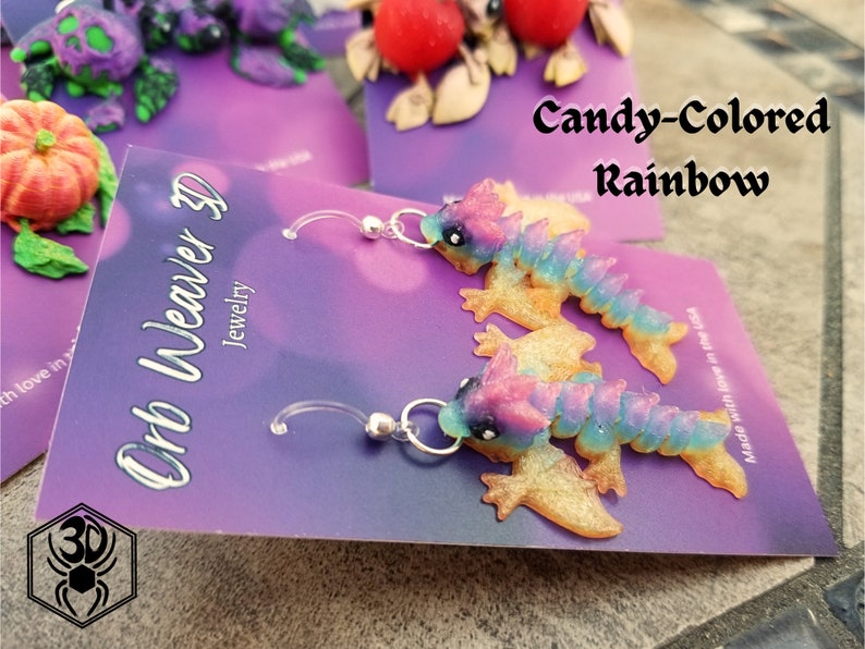 K&ouml;nnte beinhalten: Ein Paar H&auml;ngeohrringe mit einem verspielten, bonbonfarbenen Drachen-Design. Der Drache ist aus Polymer Clay gefertigt und hat eine glitzernde Oberfl&auml;che. Die Ohrringe befinden sich auf einer violetten Karte mit dem Text "Orb Weaver 3D Jewelry" und "Candy-Colored Rainbow".