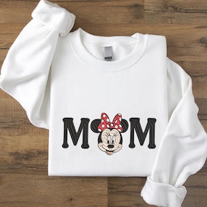 May include: White sweatshirt with the word "MOM" in black block letters, with Minnie Mouse's face replacing the "O". Minnie Mouse has a red bow with white polka dots. The sleeves are rolled up.