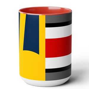 May include: A white ceramic mug with a red rim. The mug features a geometric design with yellow, blue, red, white and grey stripes.