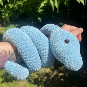 Peut inclure: Un jouet serpent en peluche bleu clair avec une tête ronde et des yeux noirs. Le serpent doux et enroulé est fait d'un matériau texturé et est tenu dans la main d'une personne sur un fond de feuillage vert.