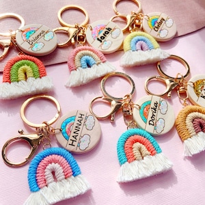 May include: A set of rainbow-themed keychains with personalized wooden tags. Each tag features a rainbow design with glitter and a name, such as Brianna, Sarah, Chelsey, Hannah, and Dorika. The keychains are made with colorful yarn and have white tassels.