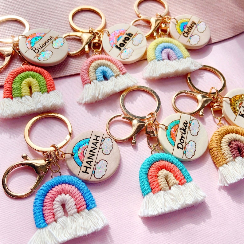 May include: A set of rainbow-themed keychains with personalized wooden tags. Each tag features a rainbow design with glitter and a name, such as Brianna, Sarah, Chelsey, Hannah, and Dorika. The keychains are made with colorful yarn and have white tassels.