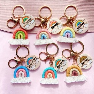 May include: Seven rainbow-themed keychains with personalized wooden tags. Each tag features a different name, including Brianna, Sarah, Chelsey, Hannah, Dorika, and Kelsey. The keychains are made with colorful yarn and have a gold-tone metal ring.