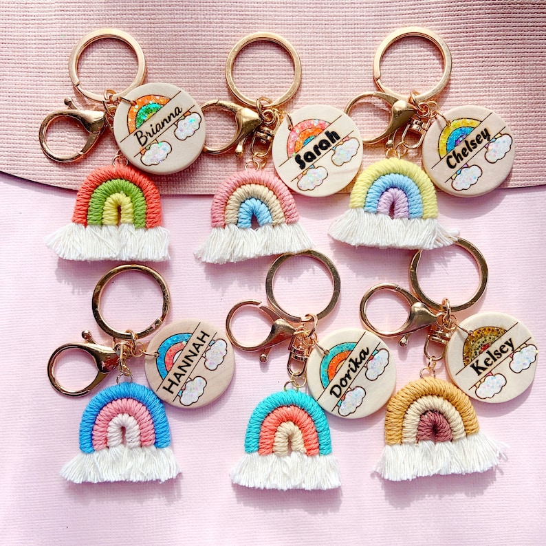 May include: Seven rainbow-themed keychains with personalized wooden tags. Each tag features a different name, including Brianna, Sarah, Chelsey, Hannah, Dorika, and Kelsey. The keychains are made with colorful yarn and have a gold-tone metal ring.