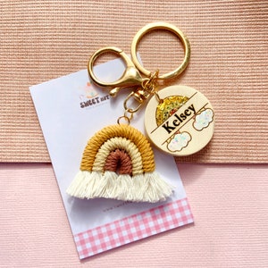 May include: A gold keychain with a rainbow-colored crocheted charm and a wooden tag with the name "Kelsey" on it. The tag has a glitter rainbow and clouds.