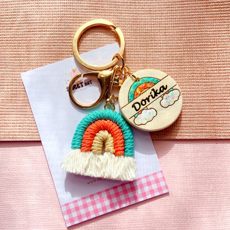 May include: A gold keychain with a rainbow-colored crocheted charm and a wooden tag with the name "DoriKa" and a rainbow design.