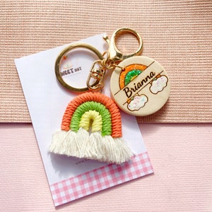 May include: A rainbow-colored keychain with a wooden charm that says "Brianna" with a rainbow and clouds. The keychain is made of yarn and has white tassels.