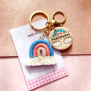 May include: A rainbow-colored crocheted keychain with white tassels attached to a gold key ring. The keychain is paired with a round wooden tag with a rainbow design and the name "HANNAH" engraved on it.