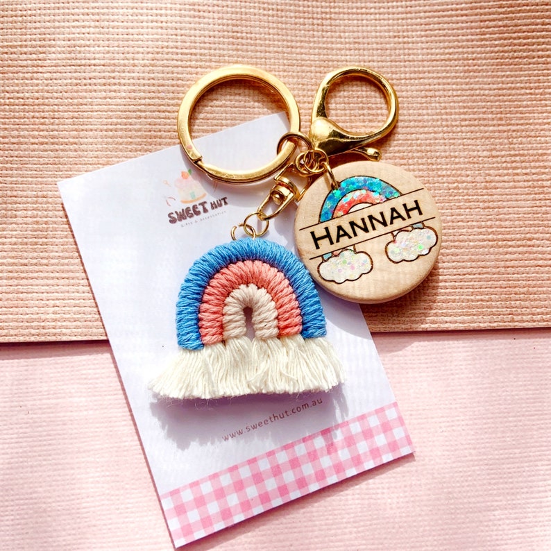 May include: A rainbow-colored crocheted keychain with white tassels attached to a gold key ring. The keychain is paired with a round wooden tag with a rainbow design and the name "HANNAH" engraved on it.